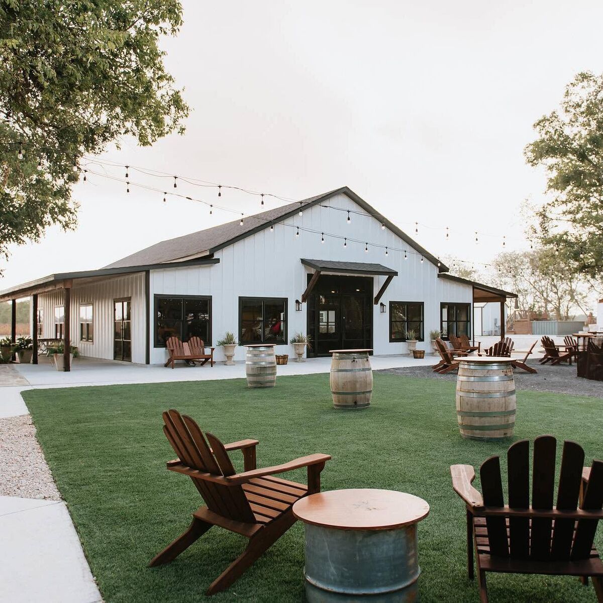 Lawn in front of the white hall with string lights, barrels and Adirondack chairs
