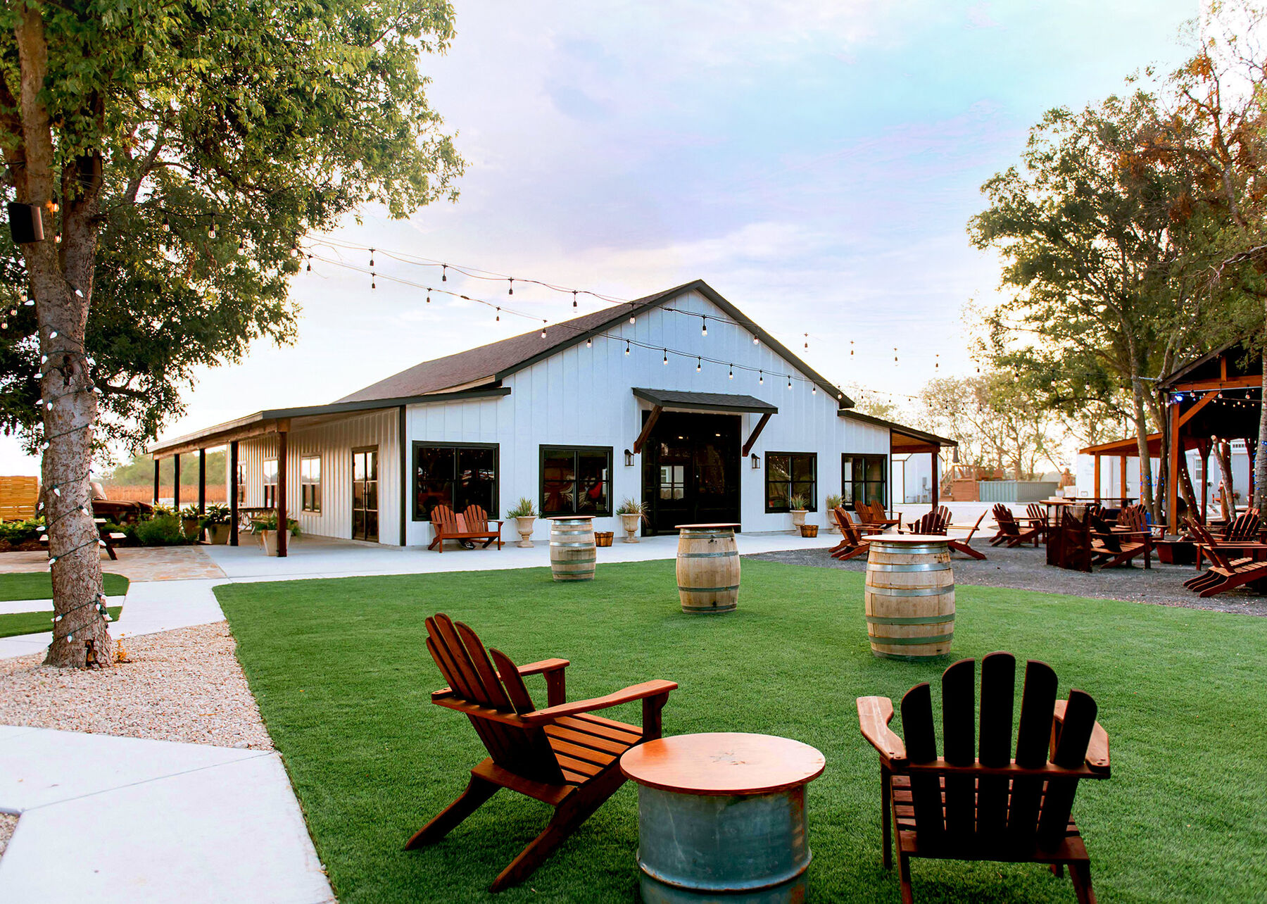 The white reception hall at The Fields with string lights, whiskey barrels and Adirondack chairs on the lawn